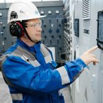Technician in safety gear working at an industrial control panel indoors.